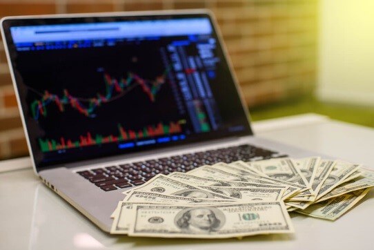 A laptop with dollar bills with a forex chart on the background – Finance Brokerage