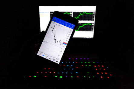 The picture displays a computer with a smartphone in the background that seems to be trading – Finance Brokerage